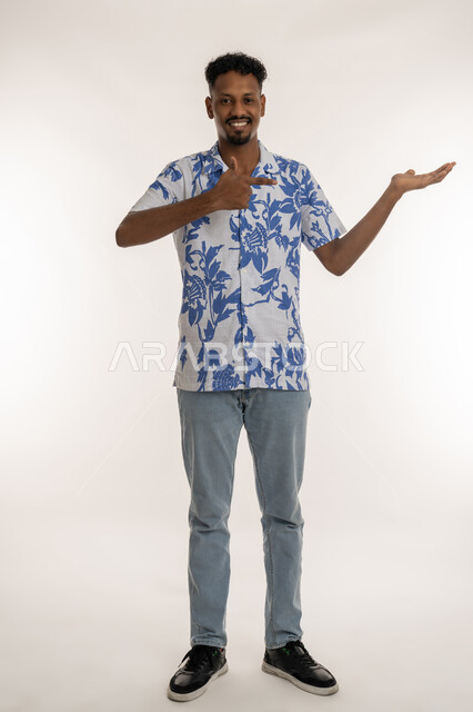 تحديد موقع شيء ما، حمل عنصر هولوجرامي، بورتريه لشاب اسمر عربي خليجي اماراتي مبتسم يرتدي الزي الكاجوال يشير الى كفه بايماءات السرور والسعادة، مفهوم التقديم والعرض، رجل سعودي ينظر الى الكاميرا بتعابير البهجة، صورة لكامل الجسم، خلفية بيضاء