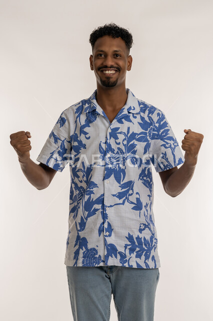 الانتصار والفوز والنجاح، مفهوم الانجاز وتحقيق الاهداف، بورتريه لشاب عربي خليجي إماراتي مبتسم يرتدي الزي الكاجوال يرفع قبضتي يديه للاعلى بإيماءات النصر، رجل سعودي ينظر لمكان ما بتعابير الفرح والسرور, الشعور بالسعادة والقوة، خلفية بيضاء