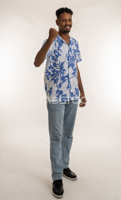 الشعور بالسعادة والقوة، الانتصار والفوز والنجاح، مفهوم الانجاز وتحقيق الاهداف، بورتريه لشاب عربي خليجي إماراتي مبتسم يرتدي الزي الكاجوال يرفع قبضته للاعلى بإيماءات النصر، رجل سعودي ينظرالى الكاميرا بتعابير الفرح والسرور, صورة لكامل الجسم، خلفية بيضاء