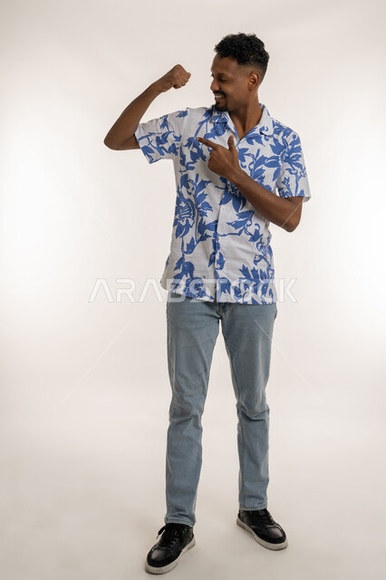 Increase body endurance, fitness concept, Saudi man exercising to get a healthy body, portrait of a young Arab Gulf Emirati man wearing casual clothes raising his hand with gestures of strength pointing his index finger to his muscles, full body image, white background