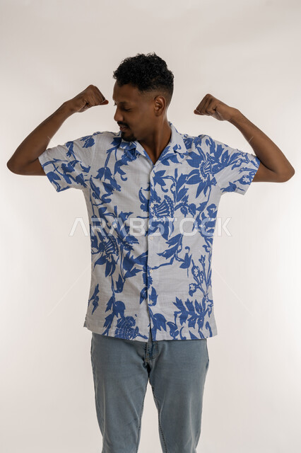 ممارسة الرياضه للحصول على جسم سليم، زيادة قدرة الجسد على التحمل، مفهوم اللياقه البدنيه، رجل سعودي يقوم بعرض العضلات ذات الرأسين، بورتريه  لشاب عربي خليجي إماراتي يرتدي الزي الكاجوال يرفع يديه بإيماءات القوة، صورة لكامل الجسم، خلفية بيضاء