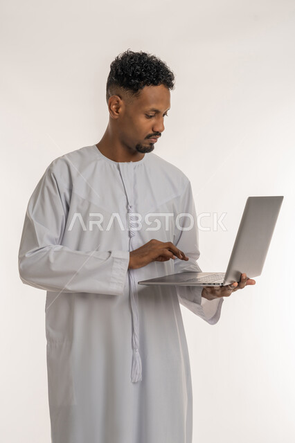 تصفح الإنترنت والعمل عن بعد، بورتريه لرجل عربي خليجي إماراتي يرتدي الكندورة الإماراتية يحمل بيده جهاز الحاسوب المحمول يقوم بالضغط على أزرار لوحة المفاتيح، استخدام الاجهزة التقنية الحديثة، إنجاز مهام العمل من خلال اللابتوب، خلفية بيضاء