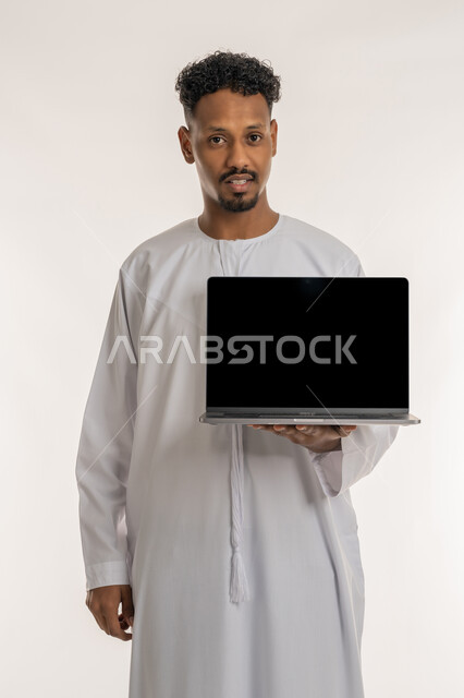 النظرالى الكاميرا بايماءات الثقة بالنفس، استخدام الاجهزة والتقنيات الحديثة، عرض صفحات الويب والبرامج والتطبيقات الالكترونية، بورتريه لرجل عربي خليجي إماراتي يرتدي الكندورة الإماراتية يحمل حاسوب محمول بشاشة سوداء فارغة، خلفية بيضاء