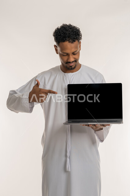 Using modern devices and technologies, displaying web pages, programs and electronic applications, portrait of an Arab Gulf Emirati man wearing the Emirati kandura holding a laptop with a blank black screen pointing his index finger towards it, white background