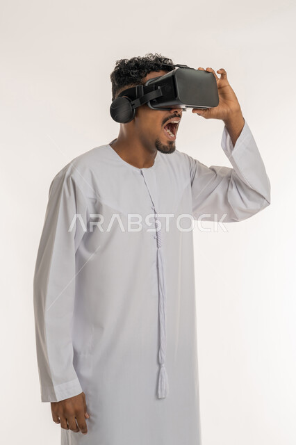 تعابير الاندهاش والتفاجؤ، المتعة و التسلية بالمشاهدة عبر نظارة الواقع الافتراضي، التطور التقني والتكنولوجي, بورتريه  لشاب عربي خليجي إماراتي يرتدي الكندورة ينظر من خلال نظارات ثلاثية الأبعاد (الفي ار), الاندماج ومحاكاة العالم الخيالي، خلفية بيضاء