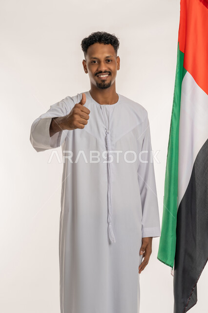 Looking at the camera with gestures of pride and honor for the country's achievements, love and belonging to the homeland, celebrating the UAE National Day, December 2, portrait of a young Arab Gulf Emirati man wearing a kandura holding the UAE flag and raising his thumb with an expression of admiration, white background