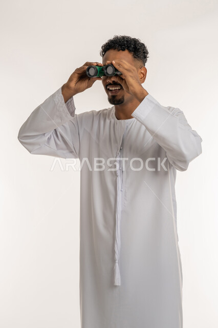 التخطيط المستقبلي للأهداف والطموحات، حب المغامرة والاستكشاف، رؤية الاشياء البعيدة، بورتريه لشاب عربي خليجي إماراتي مبتسم يرتدي الكندورة الإماراتية يحمل بيده المنظار ثنائي العينين (التلسكوب) ينظر الى الكاميرا بإيماءات السعادة والسرور والاندهاش، خلفية بيضاء