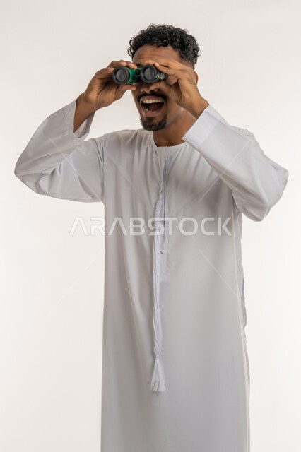 رؤية الاشياء البعيدة، بورتريه لشاب عربي خليجي إماراتي مبتسم يرتدي الكندورة الإماراتية يحمل بيده المنظار ثنائي العينين (التلسكوب) ينظر الى الكاميرا بإيماءات السعادة والسرور والاندهاش، التخطيط المستقبلي للأهداف والطموحات، حب المغامرة والاستكشاف، خلفية بيضاء