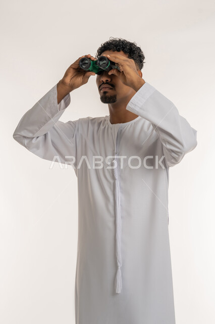 حب المغامرة والاستكشاف، رؤية الاشياء البعيدة، بورتريه لشاب عربي خليجي إماراتي مبتسم يرتدي الكندورة الإماراتية يحمل بيده المنظار ثنائي العينين (التلسكوب) ينظر الى الكاميرا بإيماءات الجدية، التخطيط المستقبلي للأهداف والطموحات، خلفية بيضاء