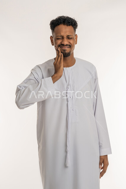 مفهوم تسوس الأضراس، الإنتفاخ والإلتهاب بالفم واللثة، ضرورة الذهاب للطبيب والحصول على العلاج, بورتريه لشاب عربي خليجي إماراتي يرتدي الكندورة يضع يده على وجهه بإيماءات الألم ووجع الأسنان، خلفية بيضاء