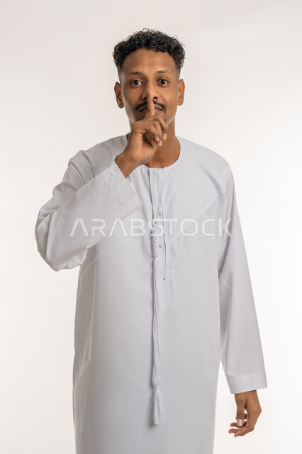 Stop talking and ask to lower the volume, order not to disturb, silence and silence, portrait of a young Arab Gulf Emirati man wearing a kandura, putting his index finger on his mouth, looking at the camera with gestures of seriousness, firmness and anger, white background