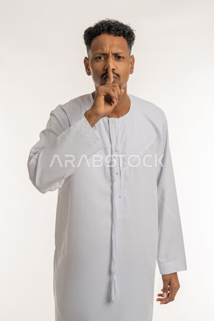 الأمر بعدم الازعاج والسكوت والصمت، بورتريه لشاب عربي خليجي إماراتي يرتدي الكندورة يضع اصبع السبابة على فمه ينظر الى الكاميرا بإيماءات الجدية والحزم والغضب، التوقف عن الكلام وطلب خفض الصوت، خلفية بيضاء