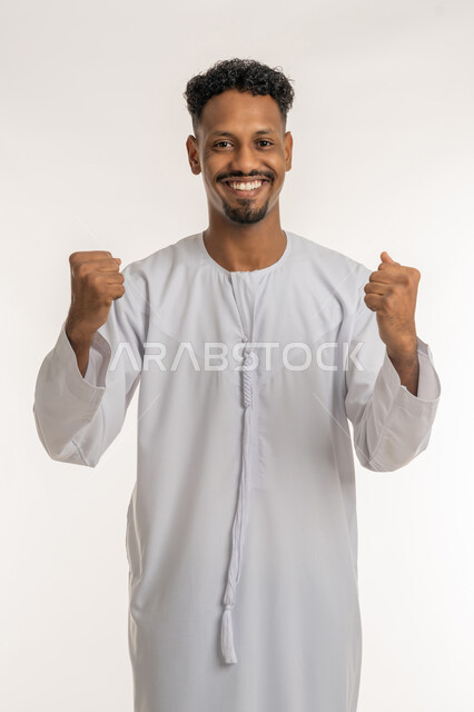 مفهوم الانجاز وتحقيق الاهداف، بورتريه لشاب عربي خليجي إماراتي مبتسم يرتدي الكندورة الإماراتية يرفع قبضتي يديه للاعلى بإيماءات الفرح والسرور, النظر إلى الكاميرا بتعابيرالسعادة والقوة، الانتصار والفوز والنجاح، خلفية بيضاء