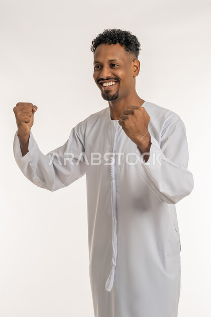 الانتصار والفوز والنجاح، مفهوم الانجاز وتحقيق الاهداف، بورتريه لشاب عربي خليجي إماراتي مبتسم يرتدي الكندورة الإماراتية يرفع قبضتي يديه للاعلى بإيماءات الفرح والسرور, الشعور بالسعادة والقوة، خلفية بيضاء