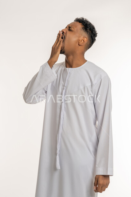 تعابير التثاؤب والخمول والرغبة في النوم، الإحساس بالإرهاق والتعب، بورتريه لشاب عربي خليجي إمارتي يرتدي الكندوره الإمارتية يضع يديه على فمه بإيماءات النعاس والحاجة الى النوم والراحة، خلفية بيضاء