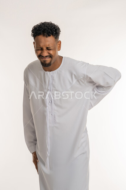 الإحساس بالألم والإصابة بمرض الديسك، بورتريه لشاب عربي خليجي إماراتي يرتدي الكندورة يقف بصعوبة ويمسك ظهره بيديه بإيماءات المعاناة والوجع، الحاجة للذهاب عند الطبيب لتلقي العلاج، آلام الظهر والعمود الفقري والتهاب المفاصل، خلفية بيضاء