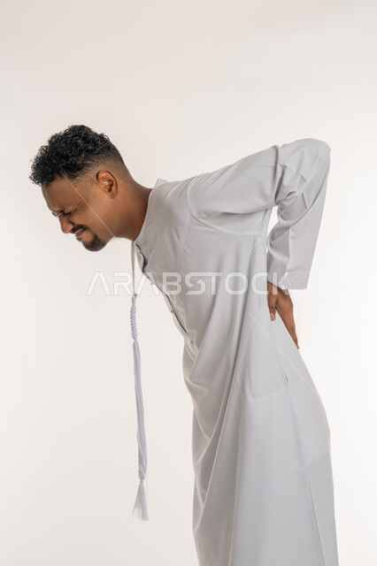 آلام الظهر والعمود الفقري والتهاب المفاصل، الإحساس بالألم والإصابة بمرض الديسك، بورتريه لشاب عربي خليجي إماراتي يرتدي الكندورة يقف بصعوبة ويمسك ظهره بيديه بإيماءات المعاناة والوجع، الحاجة للذهاب عند الطبيب لتلقي العلاج، خلفية بيضاء