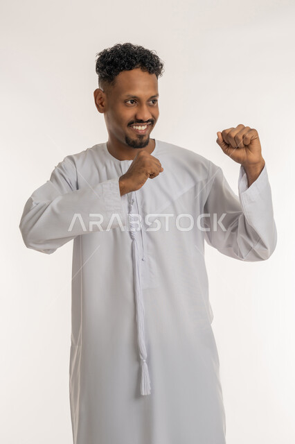 Self-defense practice, gestures of strength and challenge, mastering boxing, increasing muscle endurance, portrait of a young Arab Gulf Emirati man wearing the Emirati kandura raising his fists to block a blow, white background