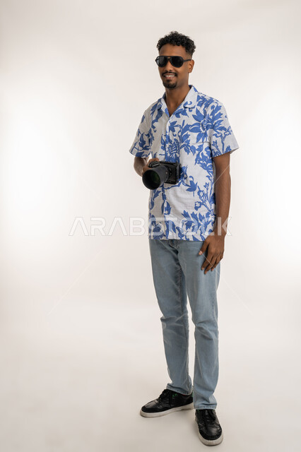 توثيق اللحظات السعيدة، إتقان واحتراف مهنة التصوير، استخدام كاميرات رقمية عالية الدقة، بورتريه لمصور محترف عربي خليجي إماراتي مبتسم يرتدي الزي الكاجوال والنظارة الشمسية، شاب سعودي يقوم بالتقاط صور تذكارية عبر كاميرا فوتوغرافية، الطول الكامل للجسم، خلفية بيضاء