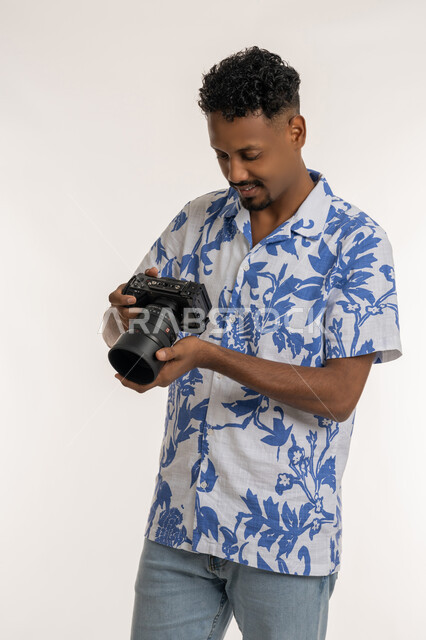 استخدام كاميرات رقمية عالية الدقة، إتقان واحتراف مهنة التصوير، بورتريه لشاب عربي خليجي إماراتي مبتسم يرتدي الزي الكاجوال يقوم بالتقاط صور تذكارية عبر كاميرا فوتوغرافية، شاب سعودي بتعابير الفرح والسرور، خلفية بيضاء