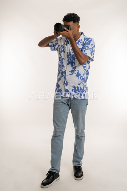 احتراف فن التصوير الفوتوجرافي باستخدام كاميرات رقمية عالية الدقة, بورتريه لمصور محترف عربي خليجي إماراتي يرتدي الزي الكاجوال، شاب سعودي يقوم بالتقاط الصور التذكارية عبر كاميرا فوتوجرافية, وظائف ومهن فنية, الطول الكامل للجسم, خلفية بيضاء