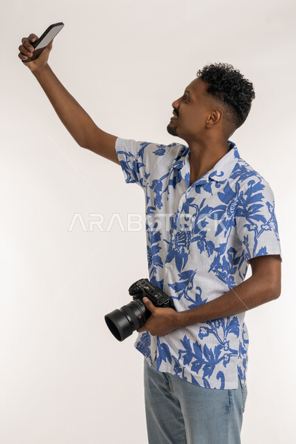 التقاط صور سيلفي من خلال الهاتف المحمول، استخدام التقنيات الحديثة المتطورة, بورتريه لشاب عربي خليجي إماراتي يرتدي الزي الكاجوال يحمل بيده كاميرا رقمية عالية الدقة، شاب سعودي يقوم بتوثيق اللحظات و الذكريات السعيدة, خلفية بيضاء