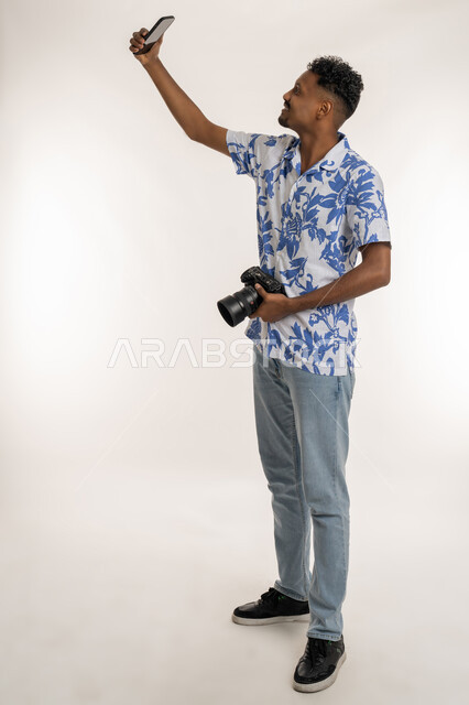 توثيق اللحظات و الذكريات السعيدة, استخدام التقنيات الحديثة المتطورة, بورتريه لشاب عربي خليجي إماراتي يرتدي الزي الكاجوال يحمل بيده كاميرا رقمية عالية الدقة، شاب سعودي يقوم بالتقاط صور سيلفي من خلال الهاتف المحمول، الطول الكامل للجسم، خلفية بيضاء