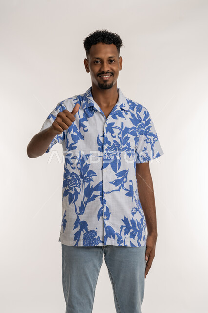 علامة الجودة والامتياز, بورتريه لشاب عربي خليجي إماراتي مبتسم يرتدي الزي الكاجوال يرفع اصبع الابهام للاعلى بإيماءات الإعجاب والرضا، مفهوم الموافقة والقبول، شاب سعودي ينظر الى الكاميرا بتعابير السعادة والسرور، النجاح وتحقيق الأهداف، خلفية بيضاء