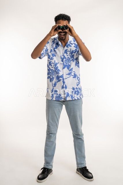 رؤية الاشياء البعيدة، بورتريه لرجل عربي خليجي إماراتي مبتسم يرتدي الزي الكاجوال يحمل بيده المنظار ثنائي العينين (التلسكوب)، شاب سعودي ينظر الى الكاميرا بإيماءات السعادة والسرور، التخطيط للأهداف والطموحات، حب المغامرة والاستكشاف، الطول الكامل للجسم، خلفية بيضاء