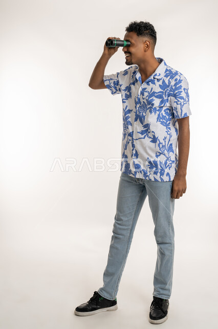 التخطيط المستقبلي للأهداف والطموحات، حب المغامرة والاستكشاف، رؤية الاشياء البعيدة، بورتريه لرجل عربي خليجي إماراتي مبتسم يرتدي الزي الكاجوال يحمل بيده المنظار ثنائي العينين (التلسكوب)، شاب سعودي ينظر الى شيء ما بإيماءات السعادة والسرور، الطول الكامل للجسم، خلفية بيضاء