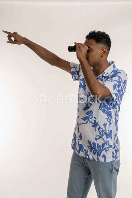 تعابير السعادة والسرور، المغامرة وحب الاستكشاف، رؤية الاشياء البعيدة، بورتريه لرجل عربي خليجي إماراتي مبتسم يرتدي الزي الكاجوال يحمل بيده المنظار ثنائي العينين (التلسكوب)، التخطيط المستقبلي للأهداف والطموحات، شاب سعودي يشير باصبع السبابة الى شيء ما،خلفية بيضاء