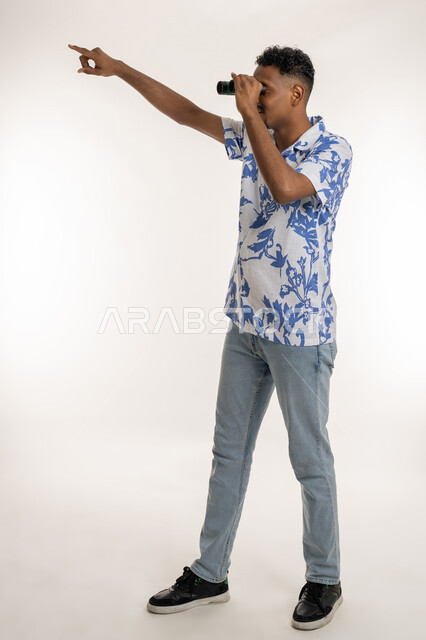 الإشارة باصبع السبابة الى شيء ما، المغامرة وحب الاستكشاف، رؤية الاشياء البعيدة، بورتريه لرجل عربي خليجي إماراتي مبتسم يرتدي الزي الكاجوال، شاب سعودي يحمل بيده المنظار ثنائي العينين (التلسكوب)، التخطيط المستقبلي للأهداف والطموحات، الطول الكامل للجسم، خلفية بيضاء
