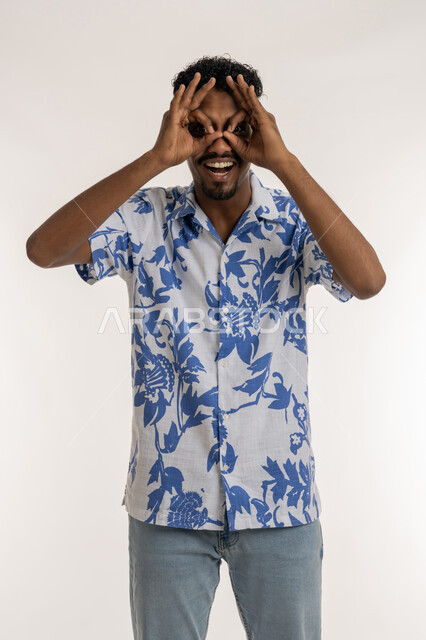 رؤية الاشياء البعيدة و تقريبها, حب الاستكشاف و المغامرة, التخطيط للأهداف والطموحات المستقبلية، بورتريه لرجل عربي خليجي إماراتي مبتسم يرتدي الزي الكاجوال، شاب سعودي يضع يديه على عينه على شكل منظار، ايماءات الاستمتاع و السرور, خلفية بيضاء