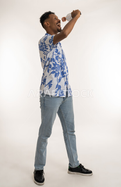 قضاء وقت ممتع بأداء العروض الصوتية، بورتريه لرجل عربي اماراتي خليجي يرتدي الزي الكاجوال يمسك بيده المايك  ويغني بحماس، شاب سعودي بايماءات الاندماج والتفاعل مع الغناء، تسجيل مقطع صوتي غنائي، تقديم الفقرات والبرامج الفنية الترفيهية، الطول الكامل للجسم، خلفية بيضاء