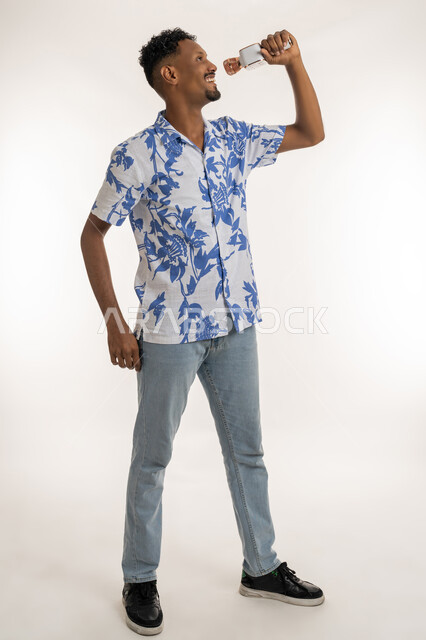 تقديم الفقرات والبرامج الفنية الترفيهية، قضاء وقت ممتع بأداء العروض الصوتية، بورتريه لرجل عربي اماراتي خليجي يرتدي الزي الكاجوال يمسك بيده المايك ويغني بحماس، شاب سعودي يسجل مقطع صوتي غنائي، ايماءات الاندماج والتفاعل مع الغناء، الطول الكامل للجسم، خلفية بيضاء