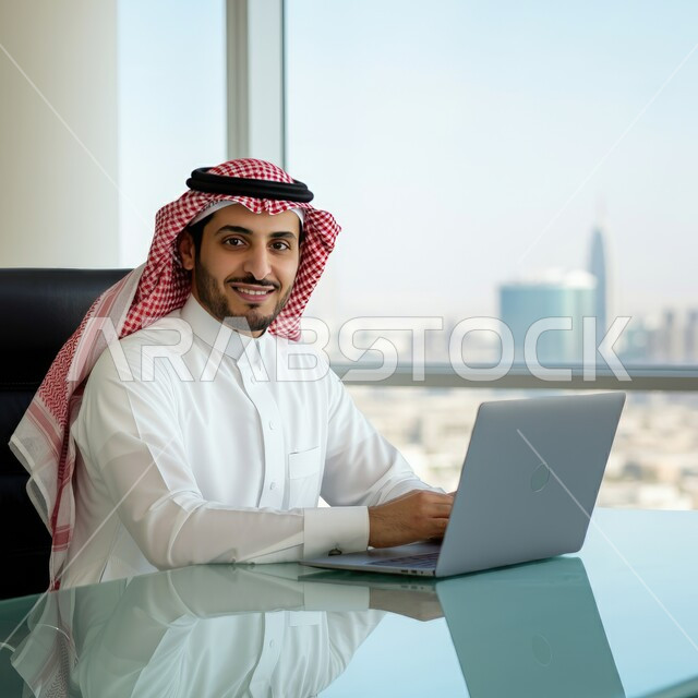 دمج التقنيات الحديثة بالقطاع الإداري والمكتبي، مهن ووظائف للشباب في السعودية، العمل في المكاتب المطلة على الأبراج وناطحات السحاب، صورة مقربة لشاب عربي خليجي سعودي يرتدي الثوب التقليدي والشماغ يجلس خلف المكتب وأمامه الحاسوب المحمول ينظر للكاميرا بإيماءات السرور