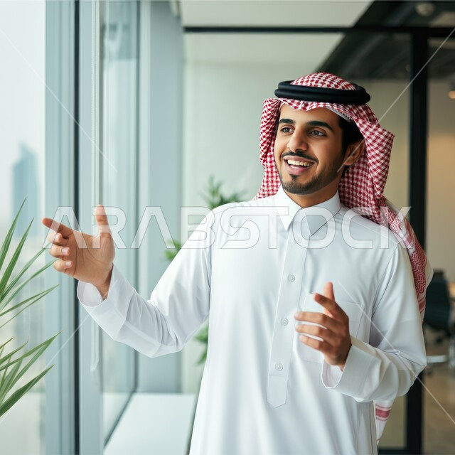 Working in offices overlooking towers and skyscrapers, employment in the administrative and office sector, professions and jobs for young people in Saudi Arabia, a close-up of a young Saudi Gulf Arab man wearing the traditional thobe and shemagh standing and looking at the urban development in the city with gestures of pleasure