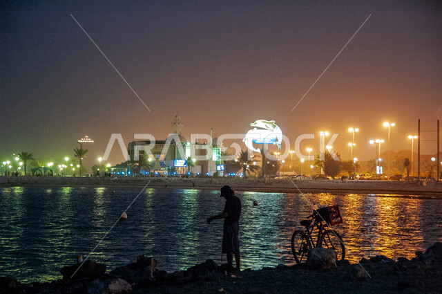 ممارسة الأنشطة الترفيهية الممتعة على الشاطئ، الفن الهندسي المعماري للمباني والمنشآت المضيئة المطلة على البحر، احتراف مهنة الصيد في المملكة العربية السعودية، رجل عربي خليجي سعودي يرتدي الزي الكاجوال يقوم باصطياد الأسماك على ساحل الخليج العربي