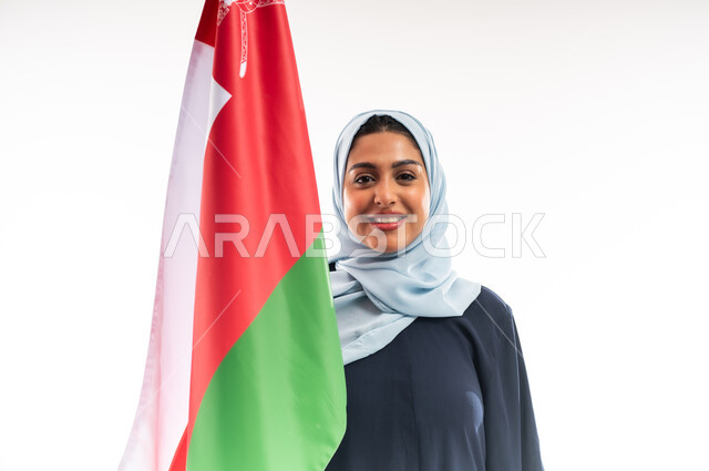 النظر الى الكاميرا بتعابير الفخر والاعتزاز، مفهوم حب الوطن، المشاركة بالاعياد والمناسبات الوطنية، اليوم الوطني العماني 18نوفمبر, احياء ذكرى استقلال الدولة 1650 م، بورتريه مقرب لشابة عربية خليجية عمانية مبتسمة ترتدي العباءة تحمل بيدها علم سلطنة عمان، خلفية بيضاء