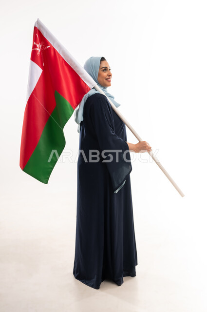 Participation in national holidays and occasions, Omani National Day November 18, the anniversary of the country's independence 1650 AD, a side portrait of a smiling young Gulf Arab Omani woman wearing an abaya carrying the flag of the Sultanate of Oman and placing it on her shoulder, pride and honor in Omani symbols, full body image, white background
