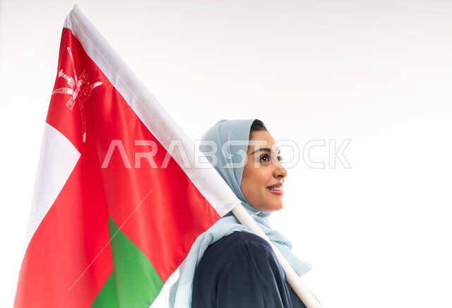 The concept of patriotism, participation in national holidays and occasions, Omani National Day November 18, the anniversary of the country's independence 1650 AD, a close-up portrait from the side of a smiling young Arab Gulf Omani woman wearing an abaya carrying the flag of the Sultanate of Oman and placing it on her shoulder, pride and honor in Omani symbols, white background