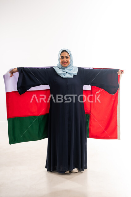 The concept of patriotism and belonging to the homeland, participation in national holidays, Omani National Day November 18, the anniversary of the country's independence 1650 AD, a portrait of a young Gulf Arab Omani woman wearing an abaya carrying the flag of the Sultanate of Oman behind her back, pride and honor in Omani symbols, a full body image, white background