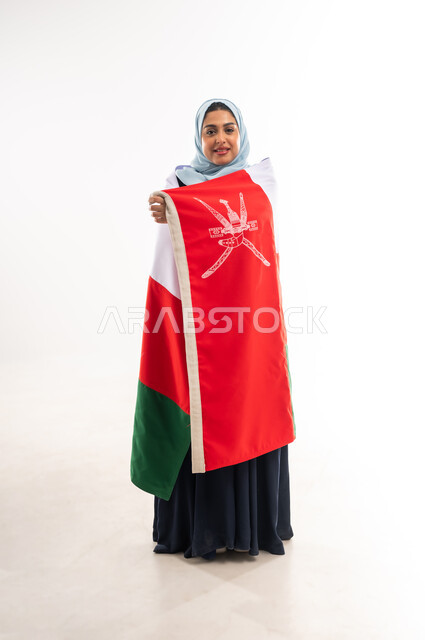 المشاركة بالاعياد والمناسبات الوطنية، الاحتفال باليوم الوطني العماني 18نوفمبر, احياء ذكرى استقلال الدولة1650م، بورتريه لشابة عربية خليجية عمانية مبتسمة ترتدي العباءة تحمل علم سلطنة عمان على ظهرها، الاعتزاز والفخر بالرموز العمانية، صورة الجسم بالكامل، خلفية بيضاء