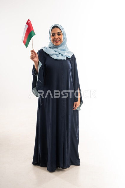 الاحتفال باليوم الوطني العماني 18نوفمبر, إحياء ذكرى استقلال الدولة 1650 م، المشاركة بالاعياد والمناسبات الوطنية، بورتريه لشابة عربية خليجية عمانية ترتدي العباءة تحمل بيدها علم سلطنة عمان، النظر الى الكاميرا بتعابير الفخر والاعتزاز، صورة الجسم بالكامل، خلفية بيضاء