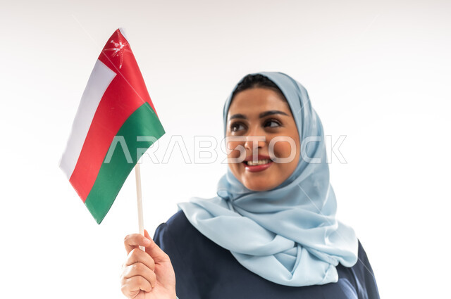 حب الوطن والانتماء اليه، الاحتفال باليوم الوطني العماني 18نوفمبر, إحياء ذكرى استقلال الدولة 1650م، المشاركة بالاعياد والمناسبات الوطنية، بورتريه مقرب لشابة عربية خليجية عمانية ترتدي العباءة تحمل بيدها علم سلطنة عمان، الفخر والاعتزاز بالرموز العمانية، خلفية بيضاء