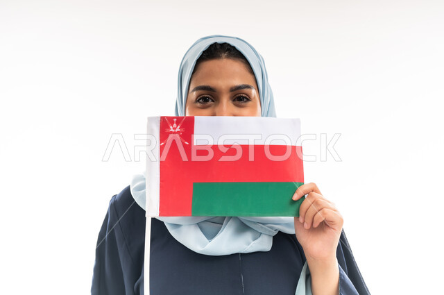 الاحتفال بذكرى اليوم الوطني العماني 18 نوفمبر, إحياء ذكرى استقلال الدولة 1650 م، المشاركة بالاعياد والمناسبات الوطنية، بورتريه مقرب لشابة عربية خليجية عمانية ترتدي العباءة تحمل بيدها علم سلطنة عمان وتضعه على وجهها، الاعتزاز والفخر بالرموز العمانية، خلفية بيضاء