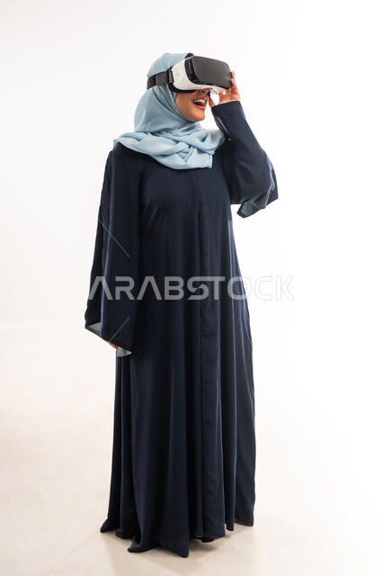 الاندماج ومحاكاة العالم الخيالي، شابة سعودية تضع يدها على نظارة الواقع الافتراضي بتعابير السعادة والسرور، بورتريه لإمرأة عربية خليجية عمانية مبتسمة ترتدي الحجاب والعباءة تنظر من خلال نظارات ثلاثية الأبعاد، التطور التقني والتكنولوجي, الطول الكامل للجسم، خلفيه يضاء