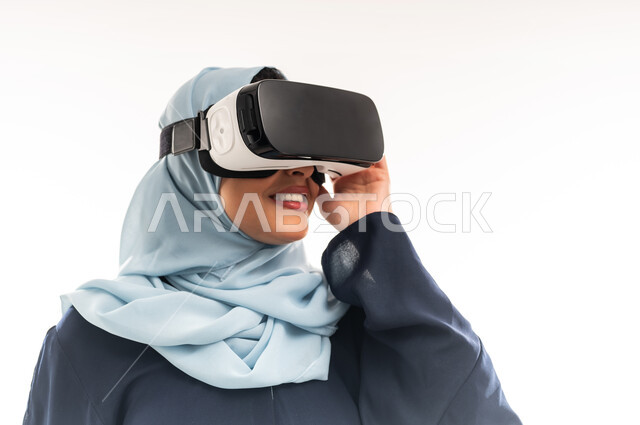الاندماج ومحاكاة العالم الخيالي، شابة سعودية تضع يديها على نظارة الواقع الافتراضي، الاستمتاع والتسلية بالمشاهدة، بورتريه مقرب لإمرأة عربية خليجية عمانية مبتسمة ترتدي الحجاب والعباءة تنظر من خلال نظارات ثلاثية الأبعاد بايماءات السعادة والسرور، خلفيه بيضاء