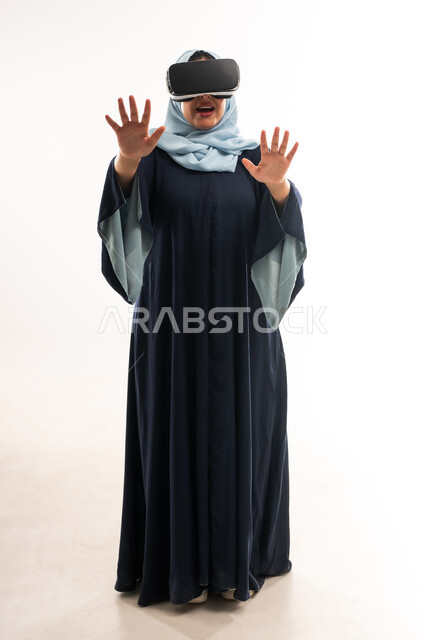 محاكاة العالم الخيالي عبر نظارات الفي آر, شابة سعودية ترفع يديها لمحاولة لمس شيء بإيماءات الاندماج، بورتريه لإمرأة عربية خليجية عمانية ترتدي العباءة تضع نظارة ثلاثية الأبعاد تقوم بالتحرك والتفاعل معها، استخدام تقنية الواقع الافتراضي, صورة الجسم بالكامل، خلفية بيضاء