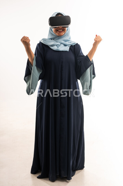 استخدام الاجهزة الحديثة والتكنولوجيا، الاندماج ومحاكاة العالم الخيالي, بورتريه لإمرأة عربية خليجية عمانية محجبة ترتدي العباءة تضع نظارة الواقع الافتراضي، شابة سعودية تغلق قبضة يديها بشدة بتعابير النصر والقوة والتشجيع، صورة الجسم بالكامل، خلفية بيضاء
